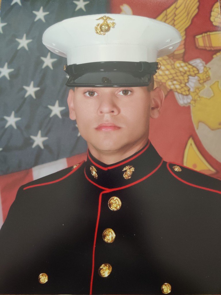 Micah Purcell in U.S. Marine Corps dress blues in front of the American flag and Marine Corps emblem.